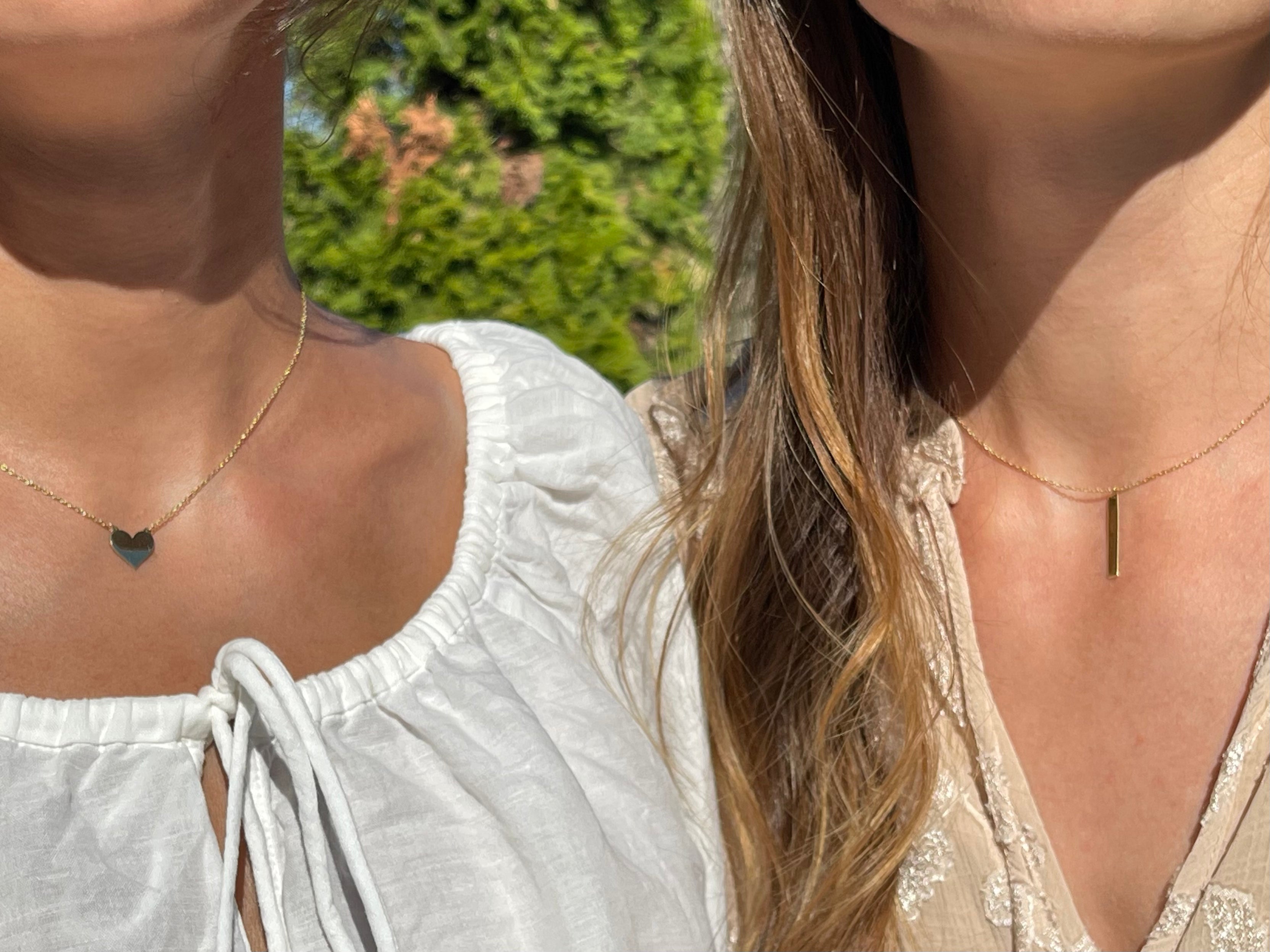 Two women wearing handmade 18k gold necklaces from Enti Jewellery, one with a heart pendant and one with a bar pendant.