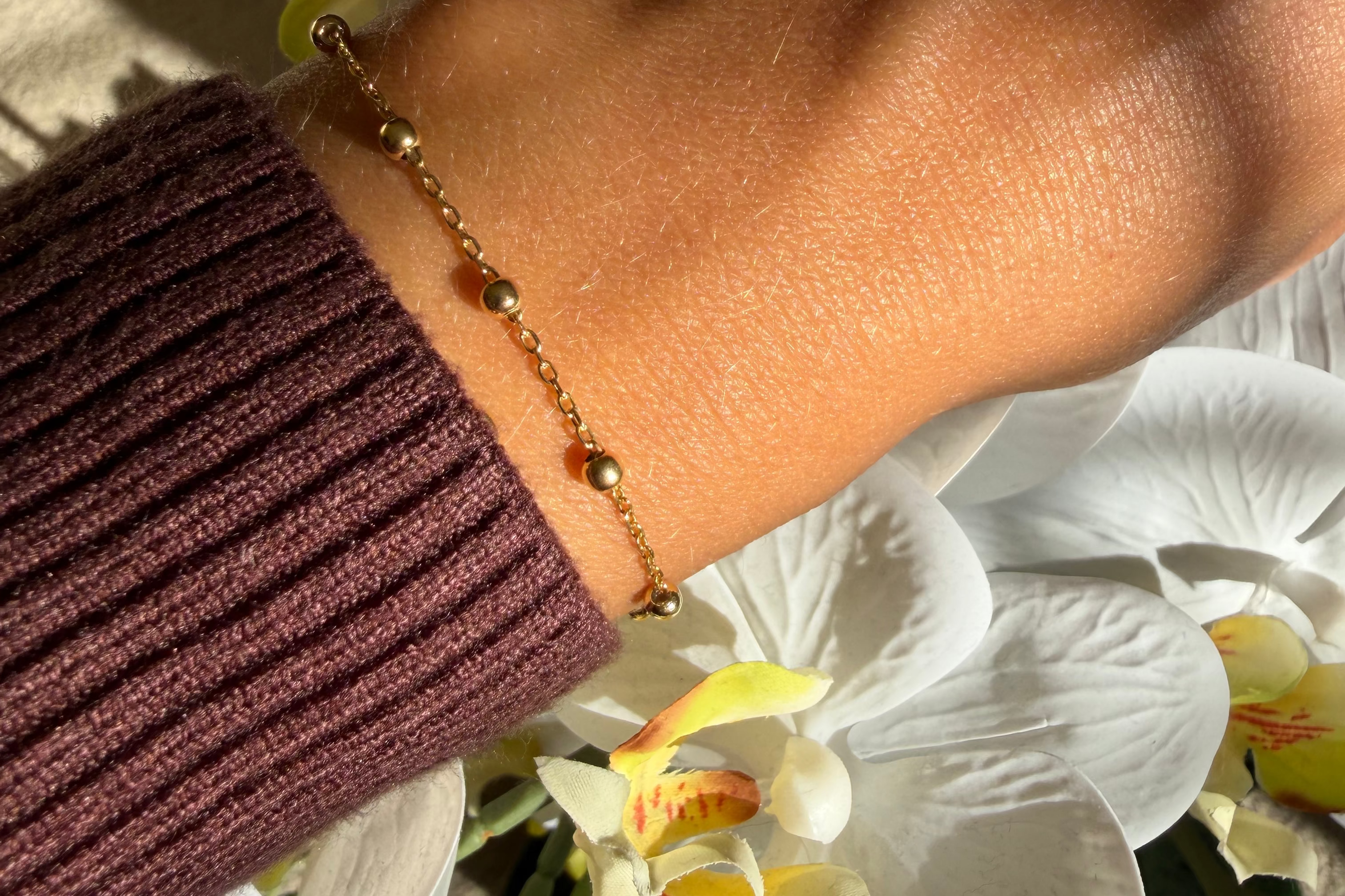A delicate gold beaded bracelet with small green bead accents worn on a person's wrist, styled with a dark burgundy ribbed sweater sleeve and white orchids in the background.